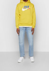 En person som bär en gul Nike-hoodie, ljusblå jeans och vita sneakers, stående mot en enkel ljusgrå bakgrund.