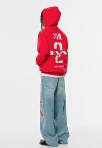 Person, der einen roten Hoodie mit weißem "EVA 2" und japanischem Text auf dem Rücken, hellblauen weiten Jeans mit rotem Seitenaufdruck und schwarzen Schuhen trägt.