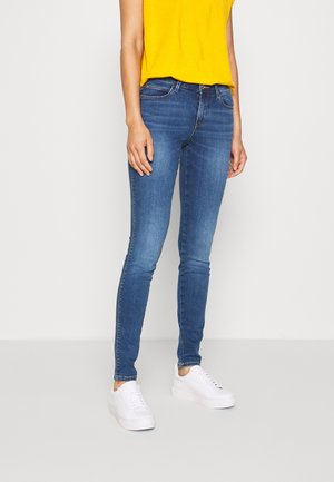 Kvinde iført tætsiddende blå jeans, hvide sneakers og en gul skjorte, stående foran en ensfarvet lys baggrund.