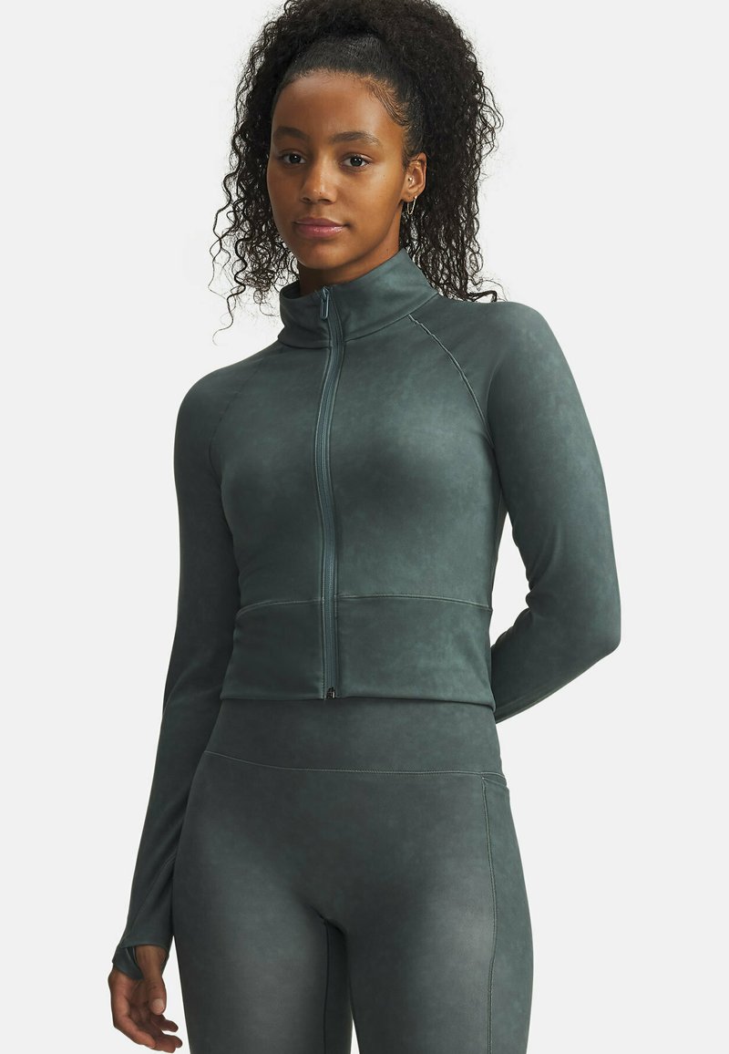 Woman with curly hair wearing dark green zip-up athletic jacket and matching leggings, standing with one arm behind back against white background.