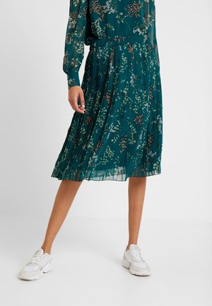 Woman in teal floral pleated skirt and matching long-sleeve top paired with white sneakers, standing against plain light background.