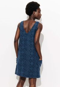 Robe bleue avec un décolleté en V, motifs circulaires et en diamant, sans manches et avec des poches latérales. Fabriquée en tissu texturé.