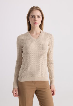 Jeune femme aux cheveux blonds lisses porte un pull beige en maille torsadée à col en V et un pantalon marron sur un fond clair uni.
