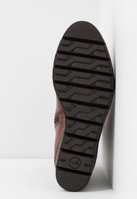 Semelle d'une chaussure en cuir marron avec des rainures profondes et un logo circulaire, positionnée verticalement contre un mur blanc.