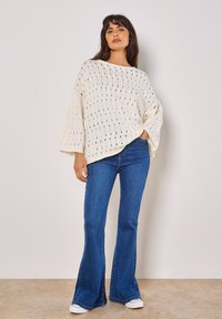 Weißer, übergroßer Strickpullover mit Lochmuster, kombiniert mit ausgestellten Jeans. Das Model steht vor einem neutralen Hintergrund, die Hände in die Hüften gestützt.