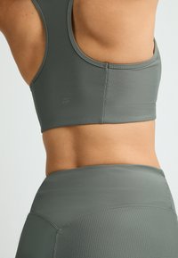 Bra sportivo a coste verde oliva con spalline larghe, caratterizzato da un design aderente e un logo discreto sul retro, abbinato a leggings a vita alta.