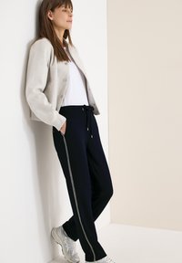 Femme appuyée contre un mur, portant une veste légère, une chemise blanche, un pantalon noir avec une bande latérale, et des baskets blanches.