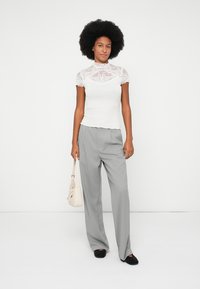 Mujer con peinado afro, vestida con una blusa de encaje blanca y pantalones gris de pierna ancha, sosteniendo un bolso crema, de pie contra un fondo blanco.