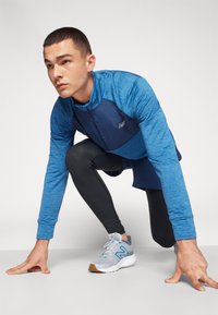 Giacca blu a maniche lunghe con zip sul davanti e pannello superiore scuro, abbinata a leggings neri e scarpe da ginnastica grigie con accenti blu.