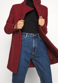 Rød uldjakke med høj krave, to knapper og en struktureret overflade. Kombineret med blå højtaljede jeans med synlige syningsdetaljer.