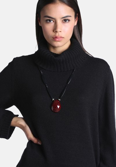 Jeune femme portant un pull à col roulé noir et un collier noir avec un grand pendentif rouge, debout, une main sur la hanche.