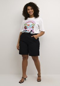 T-shirt grafica bianca con un design di auto d'epoca colorato e palme, abbinata a pantaloni corti neri fino al ginocchio. Tessuto liscio, vestibilità casual.