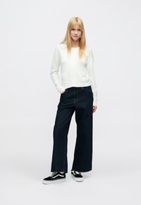 Biely kardigán s okrúhlym výstrihom a prednými vreckami, v kombinácii s tmavomodrými širokými džínsami a čiernymi teniskami.