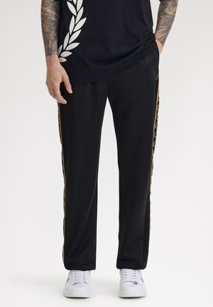 Homme portant un pantalon de survêtement noir avec une bande de laurier dorée sur les côtés et des baskets blanches, les mains dans les poches, bras tatoués visibles.