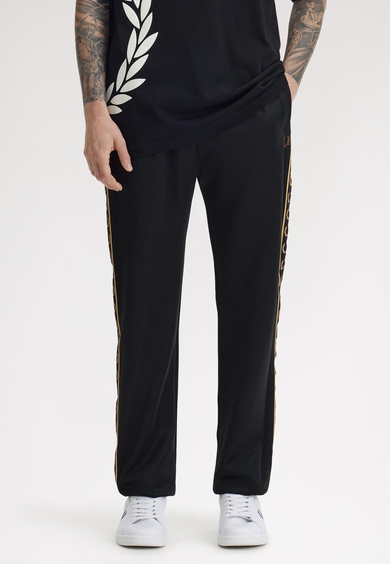 Hombre con pantalones deportivos negros con una franja dorada de laurel en los costados y zapatillas blancas, manos en los bolsillos, brazos tatuados visibles.