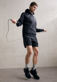 Schwarzer Hoodie mit Kängurutasche, kombiniert mit schwarzen Sportshorts. Das Modell trägt schwarze Turnschuhe und graue Crew-Socken und springt drinnen Seil.
