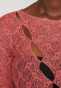 Rosa blonde topp med et mønsterdesign og utskårne detaljer. Stoffet har en teksturert finish og er lagdelt over et svart plagg under.