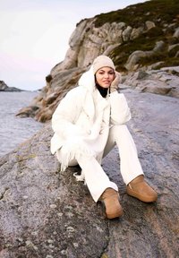 Beez värvi nahast ankle-boots kummitaldadega, valge puffer-jakk, valged püksid ja tekstuuriga mustriga kootud müts, istudes kivisel kaldal vee ääres.