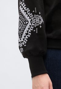 Μαύρο φούτερ με λευκό φυλετικό σχέδιο στο μανίκι, με λεπτομέρεια ριγωτής υφής στο μανσέτι και ριμπ τελείωμα.