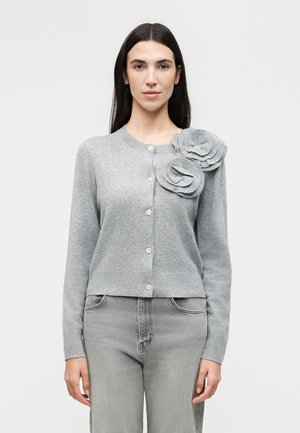 Žena nosí svetlosivý kardigán s veľkými textilnými kvetinovými ozdobami na ľavom ramene a zladené sivé džínsy.