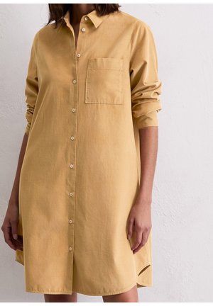 Femme portant une robe chemise boutonnée à manches longues de couleur jaune moutarde avec une poche poitrine unique, debout contre un mur blanc texturé.