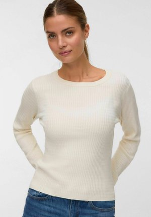 Femme à la peau claire et aux cheveux bruns attachés, portant un pull long à manches longues crème texturé et un jean bleu, devant un fond uni.