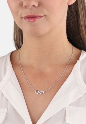 Silberne Unendlichkeitshalskette mit einer zarten Kette, die ein geschwungenes Unendlichkeitssymbol mit klaren Kristallen verziert, getragen über einer weißen Bluse.