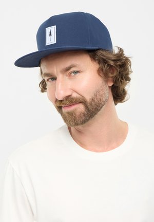 Navy blue cap with a structured brim and a white rectangular patch featuring a black tree graphic. Worn with a plain white t-shirt.