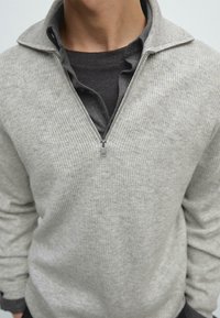 Hellgrauer Pullover mit Reißverschluss und gerippter Struktur, ausgestattet mit einem Stehkragen und über einem dunkelgrauen Hemd getragen. Der Reißverschluss ist silberfarben.