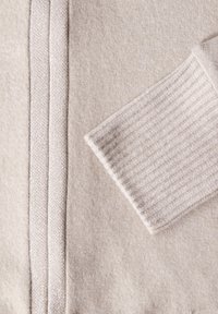 Helles beige Gewebe mit glatter Textur und subtiler glänzender Oberfläche. Verfügt über strukturierte Ärmeldetails und einen dekorativen Besatz.