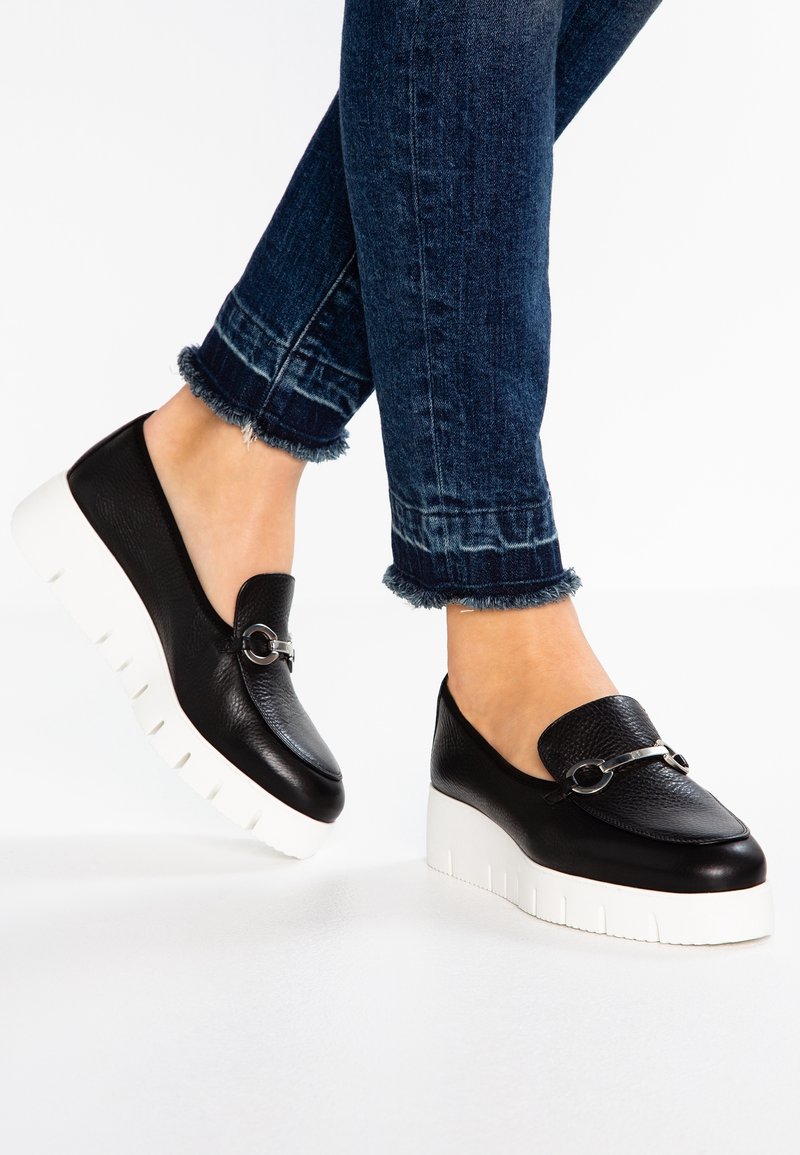Des chaussures à plateforme noires en cuir, à enfiler, avec un détail de boucle en argent, portées avec un jean bleu foncé à l'ourlet effiloché sur un sol blanc.