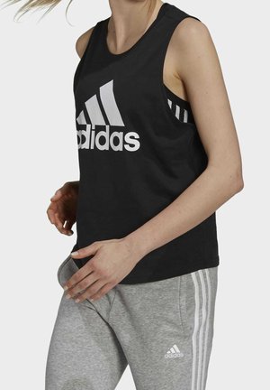 Kobieta w czarnej, bezrękawnikowej bluzce Adidas i szarych spodniach dresowych Adidas w białe paski, pozująca na jednolitym tle.
