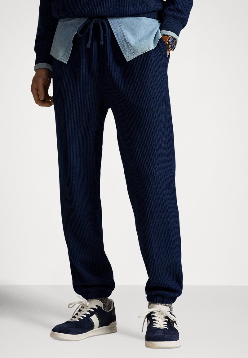 Calças de fato de treino azul-marinho com nervuras e cintura com cordão, combinadas com uma camisa de ganga azul-claro e sapatilhas escuras com detalhes brancos.