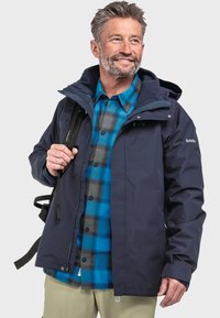Marineblauer wasserdichter Parka mit Kapuze, ausgestattet mit einem Reißverschluss. Getragen über einem blau-schwarz karierten Flanellhemd. Rucksackträger sichtbar.