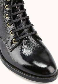 Bottine en cuir verni noir avec une finition brillante, un bout rond et six œillets dorés avec des lacets noirs. Semelle en caoutchouc avec une légère bande de roulement.