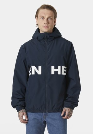 Młody mężczyzna o jasnych włosach, ubrany w granatową kurtkę z kapturem z częściowym białym napisem oraz niebieskie jeansy, stojący na jednolitym tle.