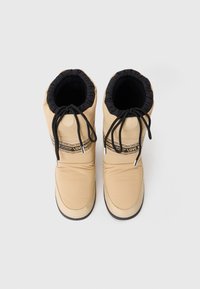 Botas isoladas bege com punho franzido, atacadores elásticos pretos e uma faixa de logótipo em contraste ao longo da parte superior. Tecido liso e almofadado.