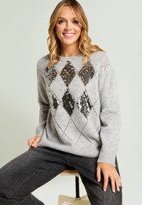 Szary sweter z diamentowym wzorem z cekinów na przodzie; długie rękawy, ściągacz u dołu i luźny krój. Ciemnoszare jeansy.