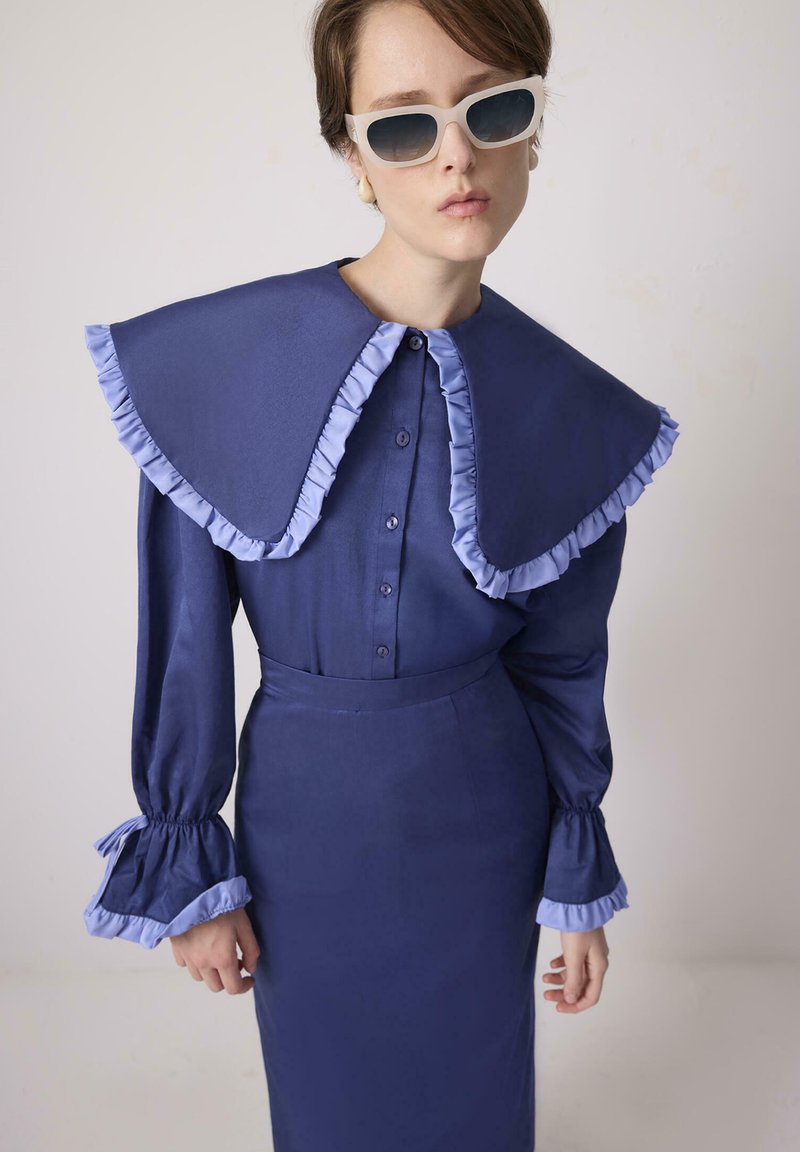 Kobieta ubrana w granatową sukienkę z dużym kołnierzem z falbanek, mankietami z falbanek oraz białymi prostokątnymi okularami przeciwsłonecznymi na jednolitym tle.