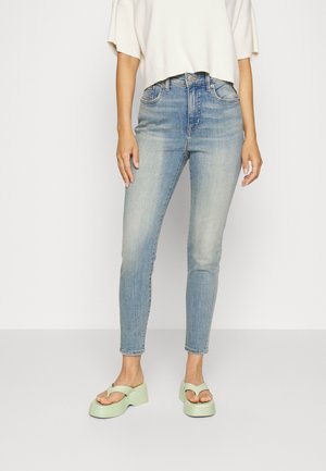 Persoon draagt lichtblauwe skinny jeans, cropped wit topje en lichtgroene platformsandalen, staand tegen een witte achtergrond.