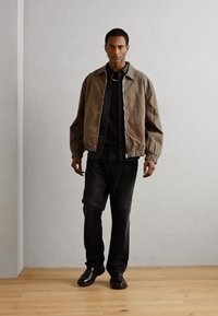 Giacca bomber marrone chiaro con zip, indossata sopra una camicia nera, abbinata a jeans scuri e scarpe nere, in piedi su un pavimento in legno.