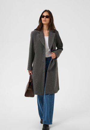 Femme portant un long manteau gris foncé par-dessus un haut blanc et un jean bleu, tenant un sac à main marron et portant des lunettes de soleil, debout devant un fond uni.