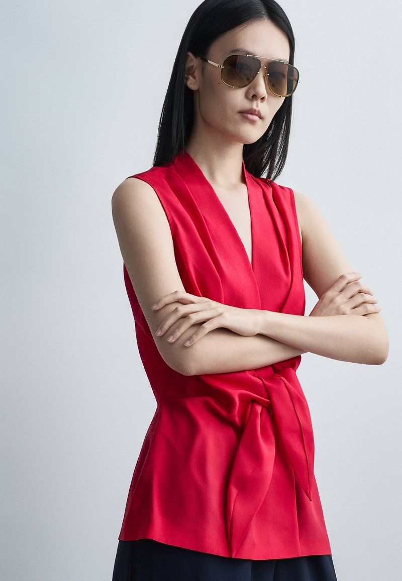 Femme aux longs cheveux noirs portant de grandes lunettes de soleil et un haut cache-cœur rouge sans manches, debout les bras croisés devant un fond uni.