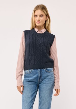 Giovane donna che indossa un gilet lavorato a trecce blu navy sopra una camicia rosa a righe con bottoni e jeans azzurri chiari, in piedi davanti a uno sfondo semplice.