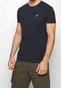 Mörkblå bomulls-T-shirt med vit rundad halsringning och liten fågel-logga på vänster bröst. Korta ärmar och standard passform.