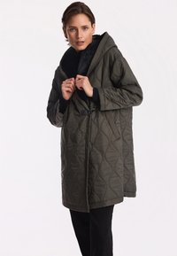 Pikowany, oliwkowozielony płaszcz z kapturem, z teksturowanym wzorem, odpinaną czarną podszewką i wcięciem w talii z detalem klamry.