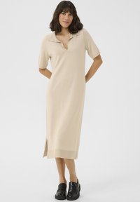 Robe midi en tricot beige avec un col, des manches courtes, une fente sur le côté et une texture douce, associée à des mocassins noirs.