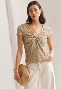 Haut smocké beige avec un décolleté en V, des manches courtes et un design de devant torsadé, associé à un pantalon taille haute de couleur claire et un sac texturé.