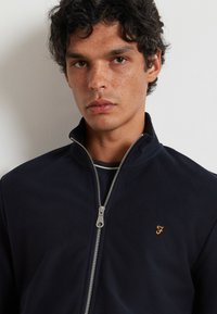 Chaqueta de cremallera azul marino con una tela texturizada, que presenta una cremallera plateada y un pequeño logo bordado en dorado en el lado izquierdo del pecho.