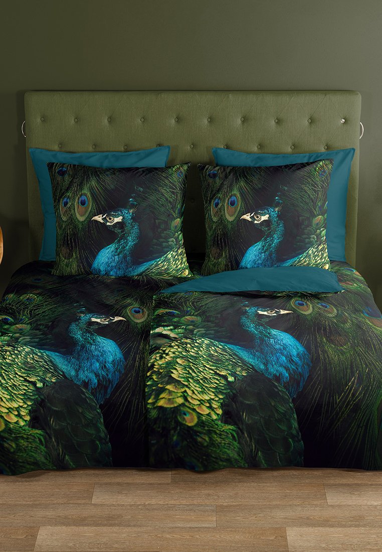 Bett mit grün gepolstertem Kopfteil, teal Kissen und Bettwäsche mit detaillierten Pfauenfeder- und blauen Pfauendekoren auf dunklem Hintergrund.
